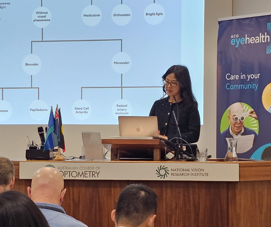 Woman with glasses delivers a lecture at a podium. Screen can be seen in background.