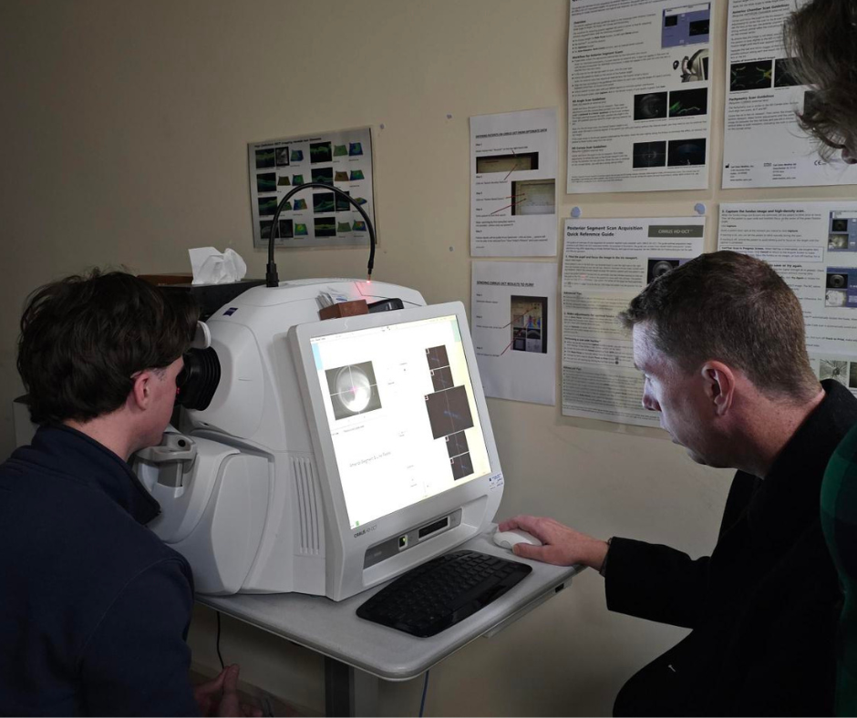 A participant sitting at screen of diagnostic machine as other participant has their eye scanned.