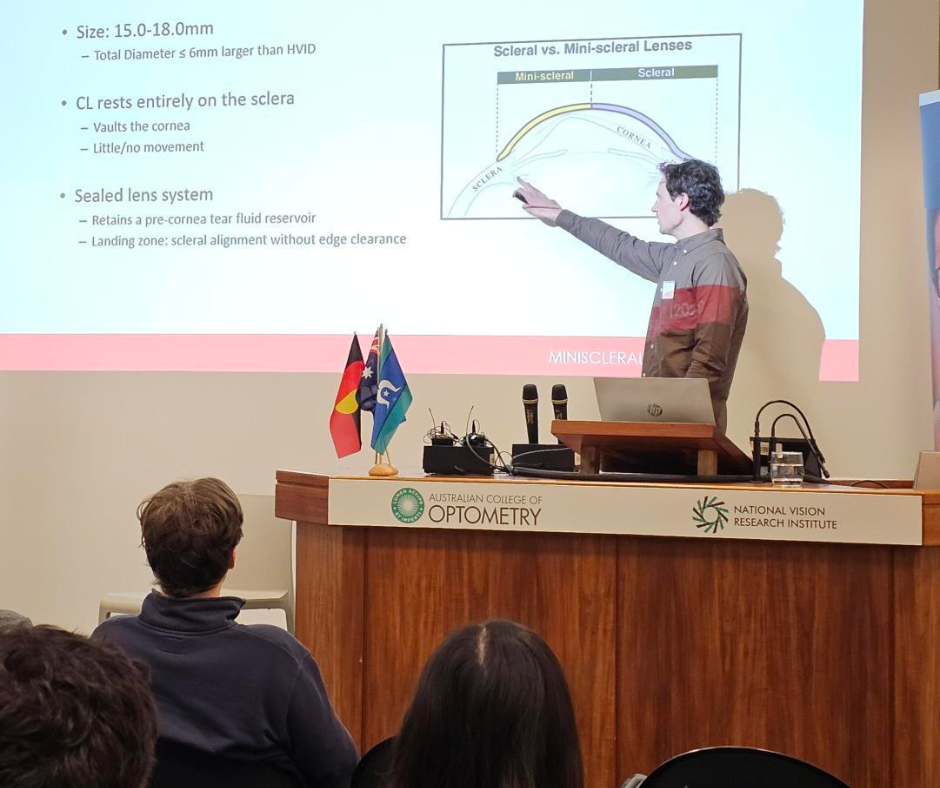 Man delivers lecture at podium, pointing to projected image on screen.