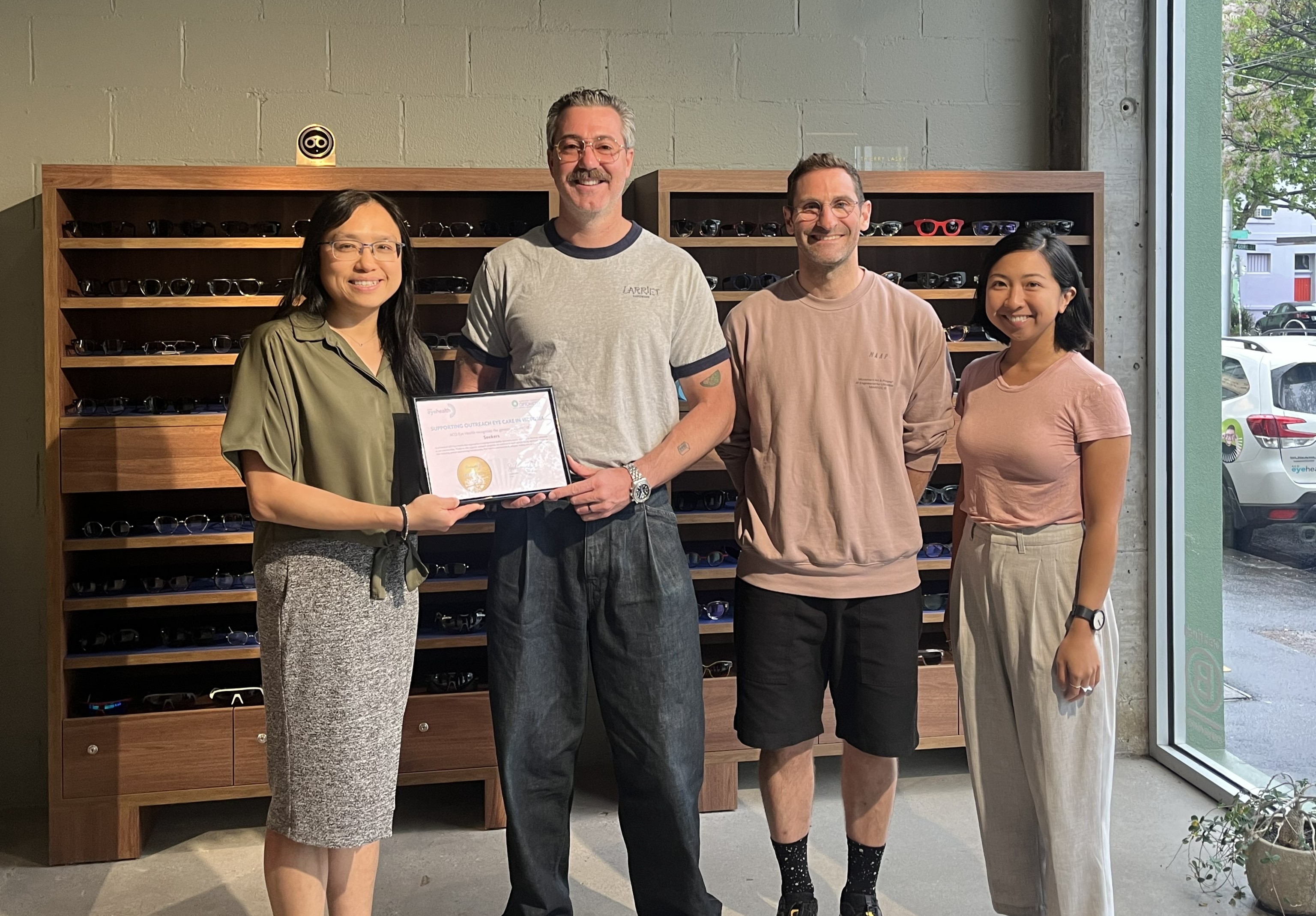 Four people stand inside an eyewear store, smiling for a photo. The person second from the left holds a certificate with a gold seal, presented by the person on the far left. Shelves filled with eyeglasses and sunglasses line the wall behind them, and a large window lets in natural light from outside.