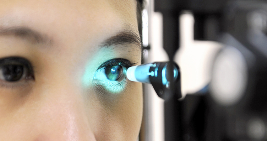 A close-up view of an eye being examined with a slit-lamp device, illuminated by a bright blue light as a clinician assesses the cornea and eye health