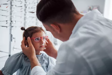 An optometrist in a white coat adjusts a pair of pink eyeglasses on a young girl. Behind them, rows of eyeglasses are neatly arranged on display racks.