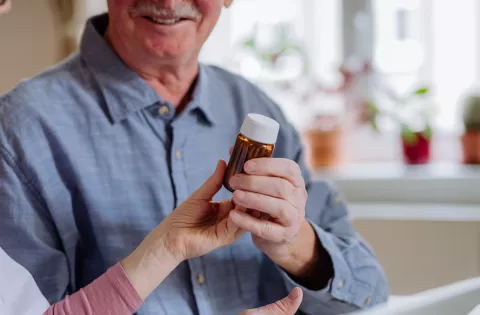 A healthcare professional is seated at a table, showing a prescription bottle to an elderly man in a blue shirt. The elderly man is smiling and engaged. The setting is a bright room with natural light and plants on the windowsill.