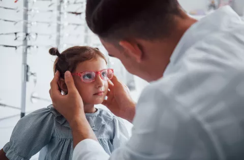 A male optometrist places a pink pair of prescription glasses on the face on a young girl, aged approximately 6 years.
