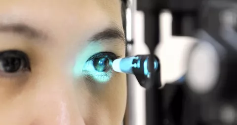 A close-up view of an eye being examined with a slit-lamp device, illuminated by a bright blue light as a clinician assesses the cornea and eye health