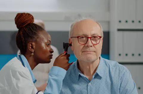 A healthcare professional in a white coat examines an older adult’s ear with an otoscope. The older adult, wearing red glasses and a light blue shirt, sits calmly in a medical office 